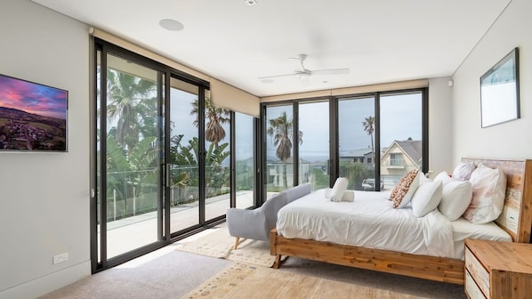 A bedroom with a large bed, a chair, and a view of palm trees and houses through large windows. - Clifftop Luxury With Pool ~ Ultimate outdoor experience with a private pool (Kiama)