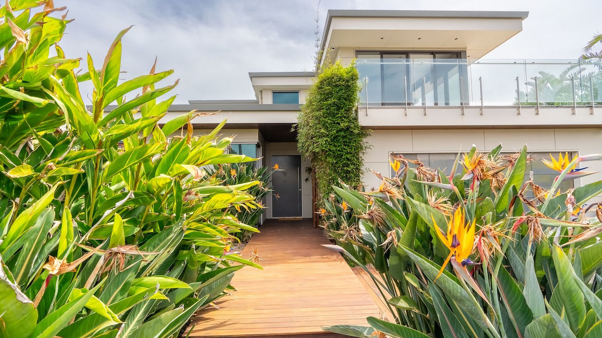 Une maison moderne dotée d’une terrasse en bois qui mène à une porte vitrée, le tout entouré d’une végétation tropicale luxuriante.