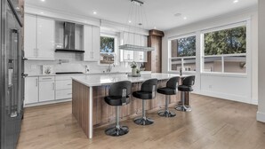 A modern kitchen with white cabinets, a large island with bar stools, and a view of the outdoors through large windows. - Luxury Spacious House (Surrey)