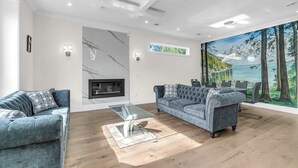 A modern living room with two sofas, a glass coffee table, a fireplace, and a large mural of a forest scene. - Luxury Spacious House (Surrey)