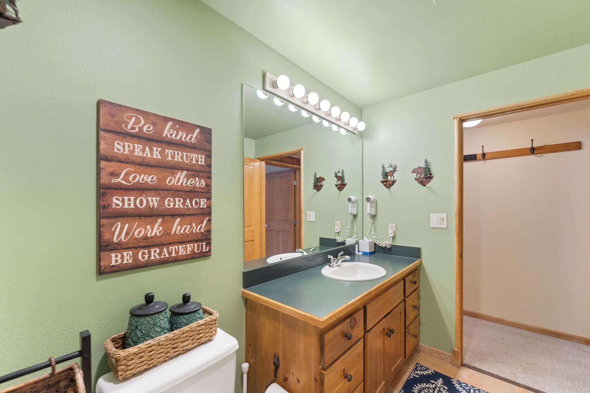 A bathroom with a wooden vanity, a mirror with lights, a sink, a toilet, and a motivational quote on the wall.