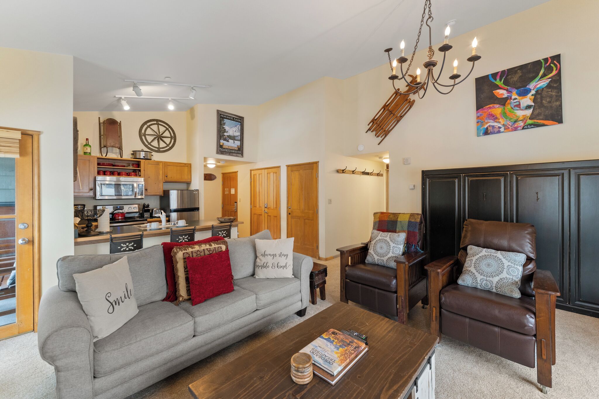 A spacious living room with a grey sofa, a brown leather chair, and a wooden coffee table. A kitchen is visible in the background with wooden cabinets and a stove.