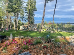 A garden with a wooden deck, surrounded by trees and a view of a lake.