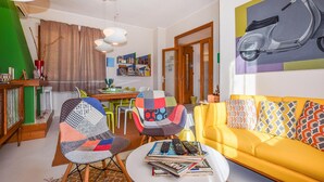 Ein Wohnzimmer mit einem gelben Sofa, bunten Kissen, einem Couchtisch mit Zeitschriften, zwei gemusterten Sessel, einem Esstisch mit Stühlen, einer grünen Wand und einem Bild von einem Roller.