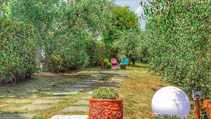 Ein Gartenpfad mit einem Blumentopf, einem weißen, kugelförmigen Objekt und bunten Stühlen im Hintergrund.