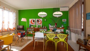 Un comedor con pared verde, una mesa de madera y sillas. Hay dos lámparas colgantes, un reloj y un florero con plantas.