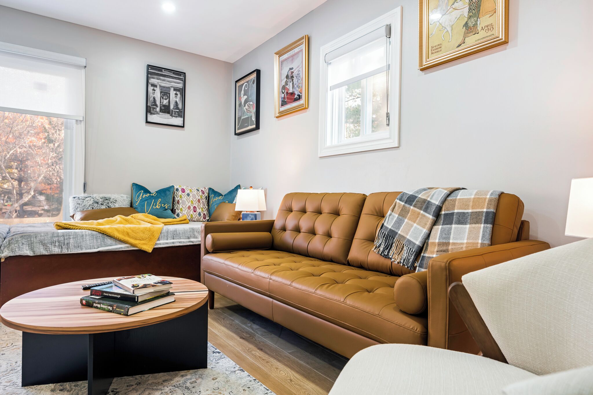 Ein Wohnzimmer mit einem braunen Sofa, einem kleinen runden Tisch mit Büchern, einem Fenster mit Jalousien und gerahmten Bildern an der Wand.