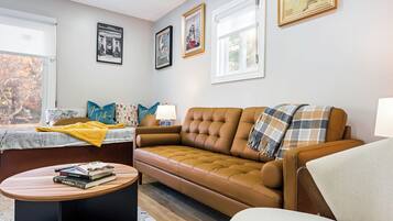 Ein Wohnzimmer mit einem braunen Sofa, einem kleinen runden Tisch mit Büchern, einem Fenster mit Jalousien und gerahmten Bildern an der Wand.