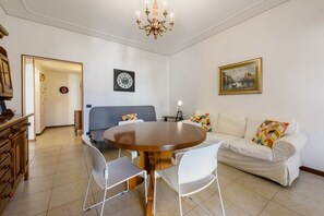 A dining area with a round wooden table, white chairs, a sofa with colorful cushions, a clock on the wall, and a chandelier. - Santa Croce's Vintage by MMega (Firenze)