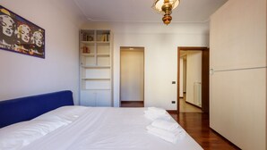 Una habitación de hotel con una cama grande, una estantería, una puerta que da a otra habitación y un pasillo.