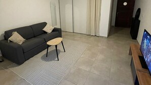 Una sala de estar moderna con un sofá gris oscuro, una pequeña mesa redonda de centro, un televisor de pantalla plana sobre un soporte de madera, y una puerta corredera que da a otra habitación.