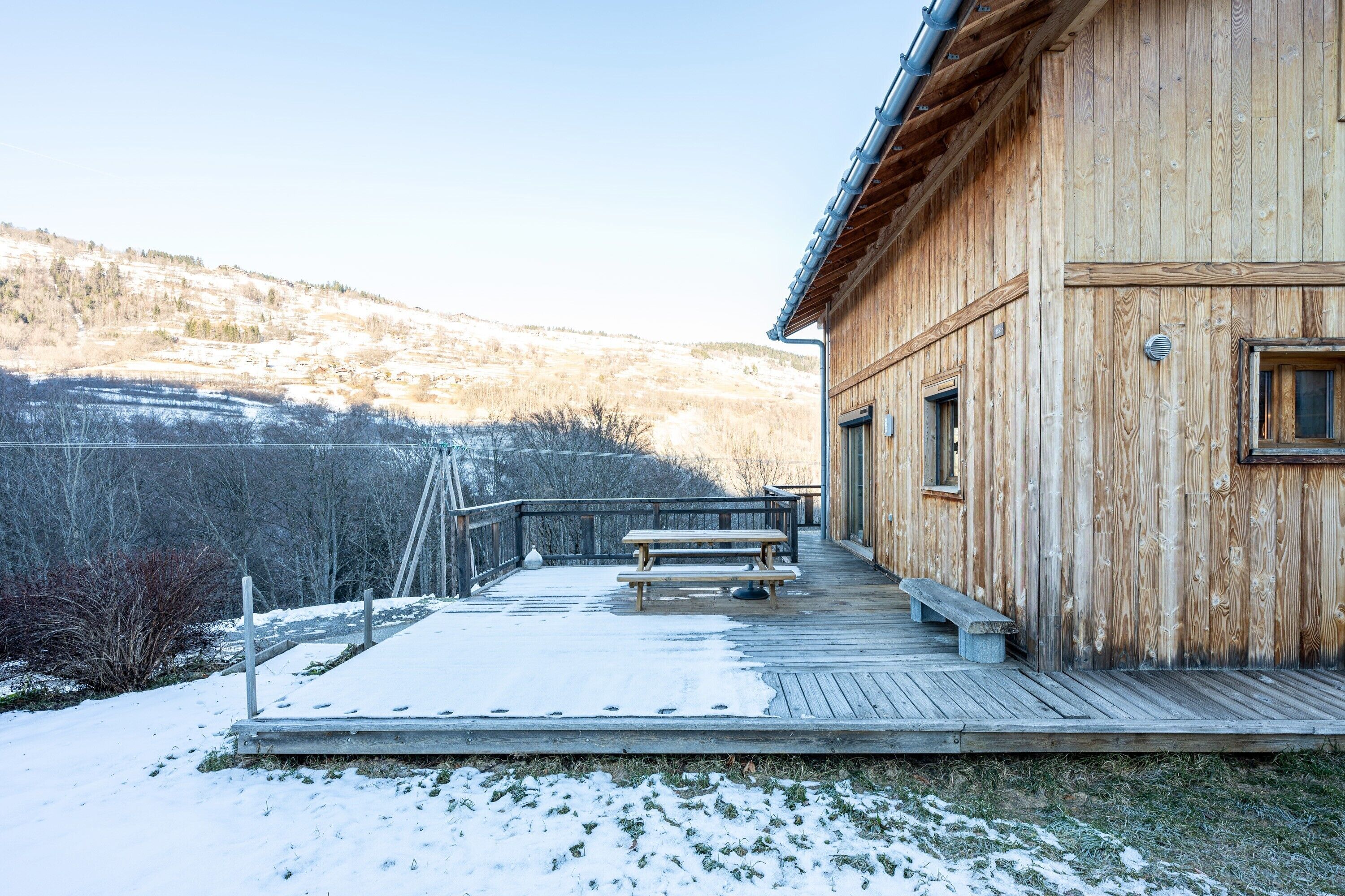 Una cabaña de madera con una terraza, un banco y vistas a una ladera nevada.