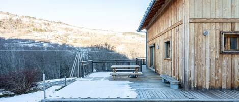Una cabaña de madera con una terraza, un banco y vistas a una ladera nevada.