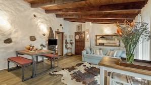 Um interior rústico com vigas de madeira expostas, parede de pedra, área de refeições com bancos, sofá e um vaso com flores.