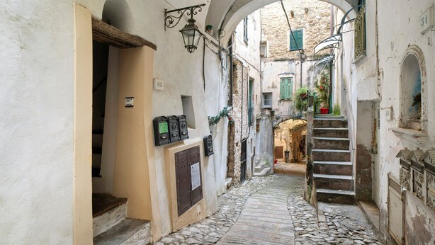 A narrow cobblestone alleyway with stone buildings, an archway, and a staircase leading to a higher level.