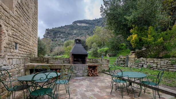 Un edificio de piedra con chimenea, asientos exteriores y vistas a una colina boscosa.