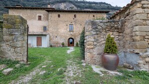 Un edificio de piedra con un portal central, un pequeño jardín con un árbol en maceta y montañas al fondo.