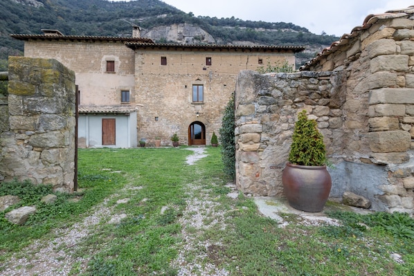 Un bâtiment en pierre avec une porte d’entrée centrale, un petit jardin agrémenté d’un arbre en pot et un paysage montagneux en arrière-plan.