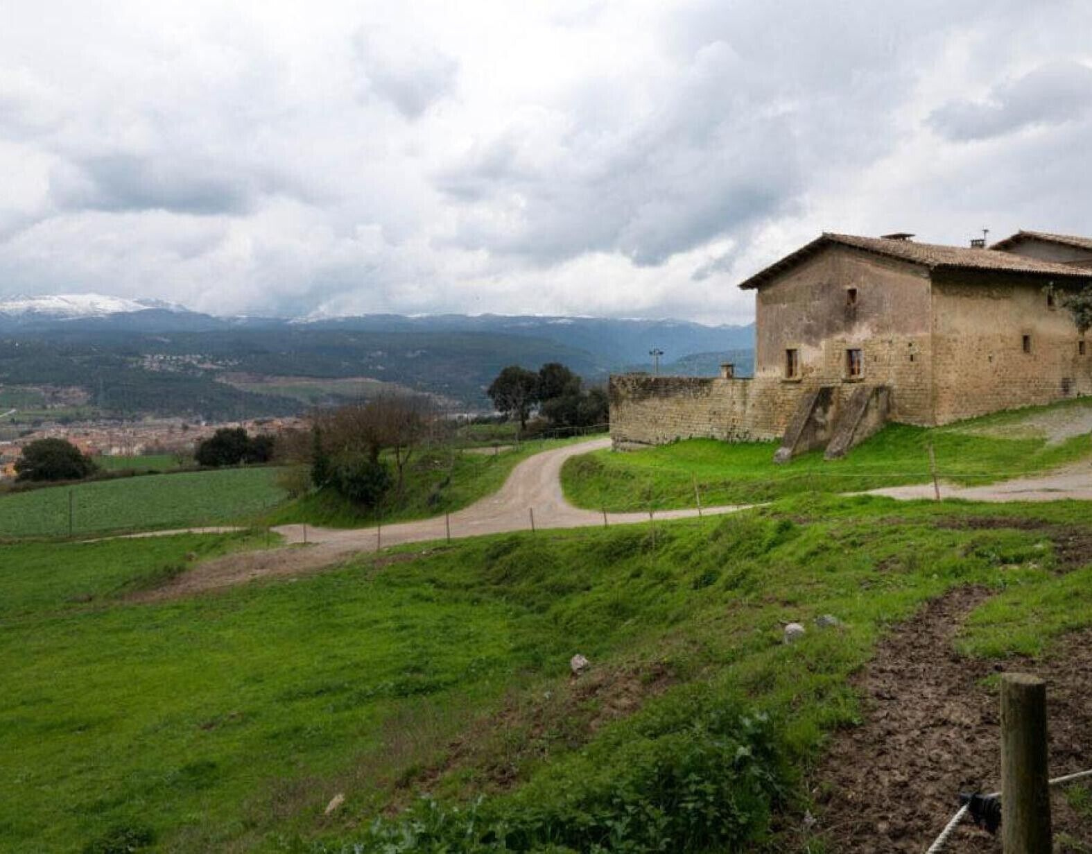 Un bâtiment en pierre doté d’un toit en pente, entouré d’une zone délimitée par une clôture et bordé d’un chemin de terre.