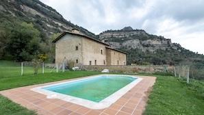 Un edificio de piedra con piscina, rodeado de césped y montañas al fondo.