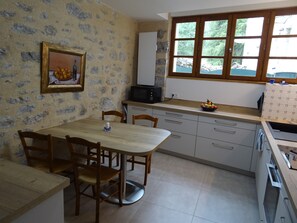 Een keuken met een eettafel, stoelen, een magnetron en een raam met uitzicht op groen.