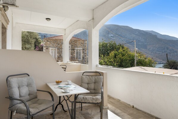 A balcony with a table and chairs, a view of mountains, and a building with a tiled roof.