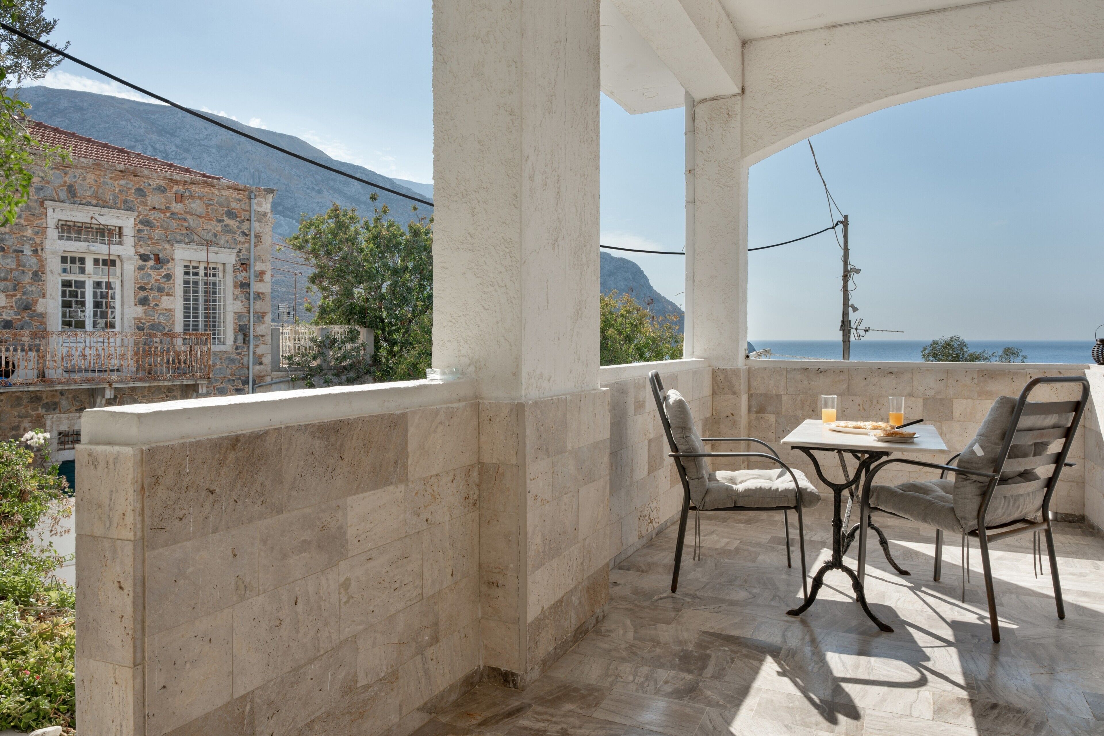 A balcony with stone walls, a table with chairs, and a view of the sea and mountains.
