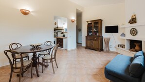 Ein Essbereich mit rundem Tisch und Stühlen, eine Küche im Hintergrund, eine Schrank mit Glastüren, ein flachbildfernseher an der Wand und ein blaues Sofa im Vordergrund.