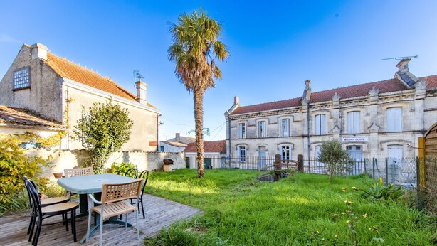 Une cour dotée d’une terrasse en bois, d’une table avec des chaises, d’un palmier et de bâtiments historiques aux façades en pierre et aux toits couverts de tuiles rouges.