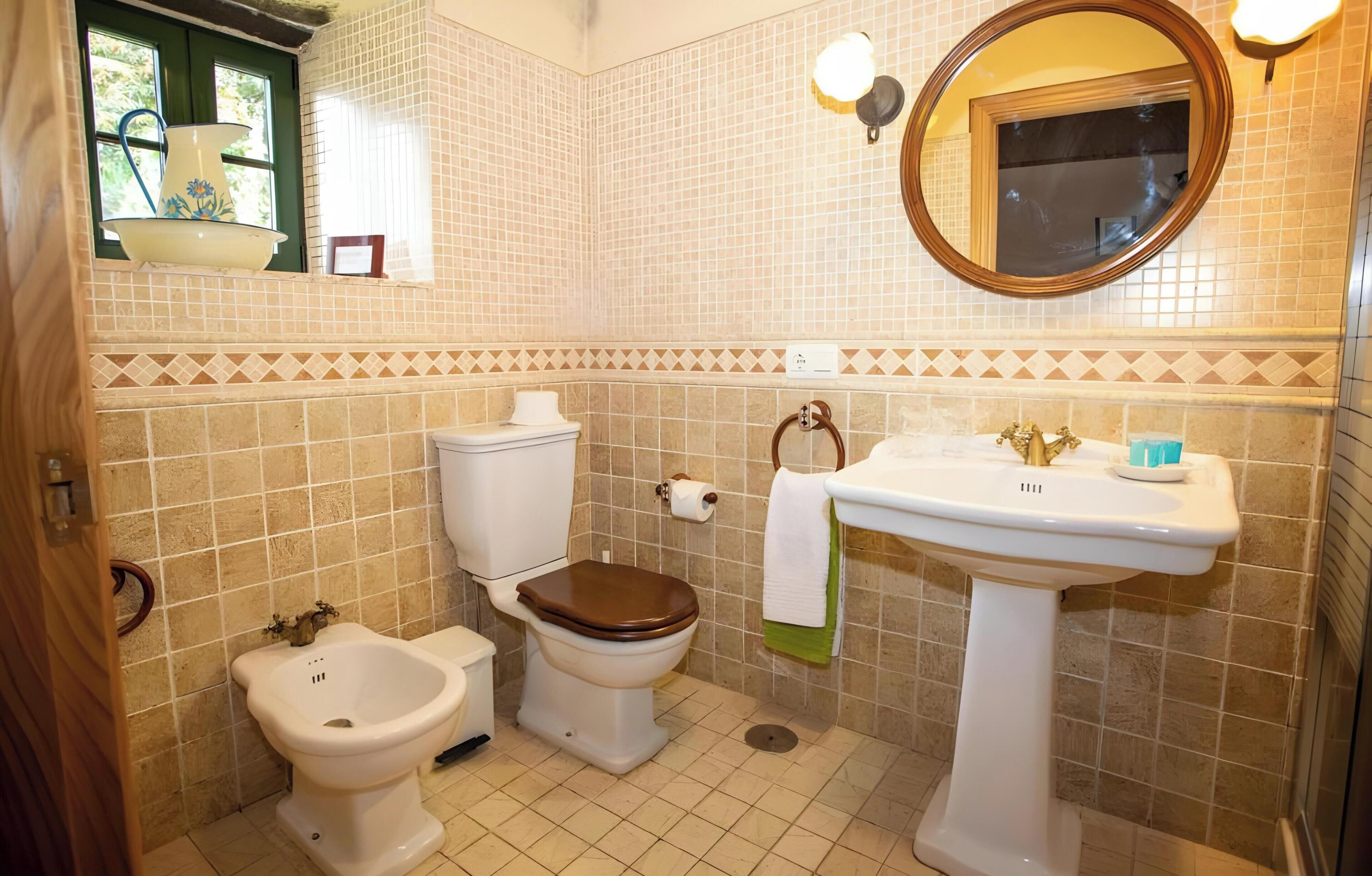 A bathroom with a toilet, bidet, sink, mirror, and tiled walls.
