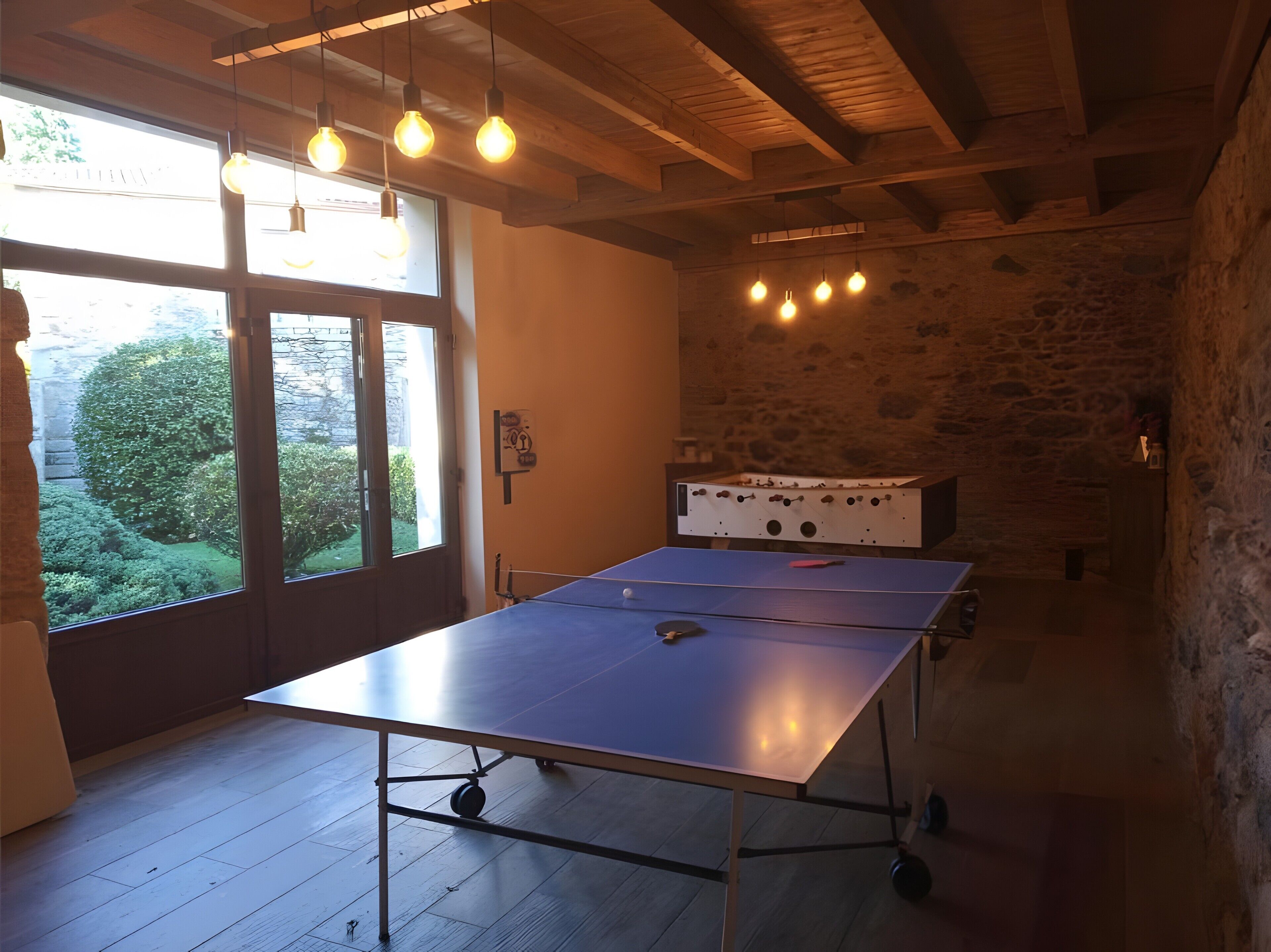 Une table de ping-pong dans une pièce aux murs de pierre, au plafond en bois et à la fenêtre donnant sur un paysage verdoyant.