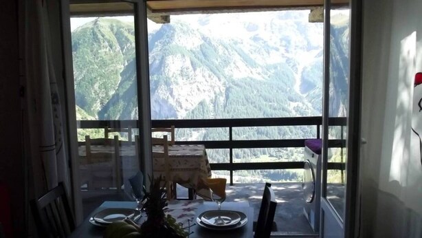 Un coin repas avec une table dressée pour deux personnes, une chaise et une vue sur les montagnes, visible à travers une porte vitrée.