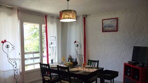 Un coin repas avec une table dressée pour quatre personnes, des chaises, un luminaire suspendu, un téléviseur posé sur un meuble et une fenêtre avec des rideaux.