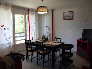 Un coin repas avec une table dressée pour quatre personnes, des chaises, un luminaire suspendu, un téléviseur posé sur un meuble et une fenêtre avec des rideaux.