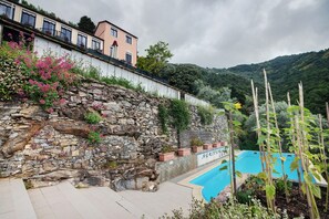 Un muro di sostegno in pietra con una piscina, un edificio con un balcone e montagne sullo sfondo.