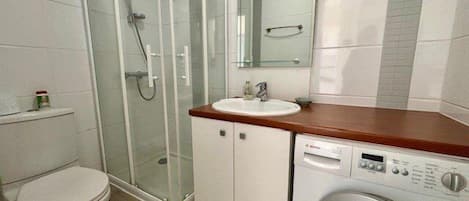 Et badeværelse med en glasafskærmet brusekabine, toilet, vask med spejl og en frontbetjent vaskemaskine.