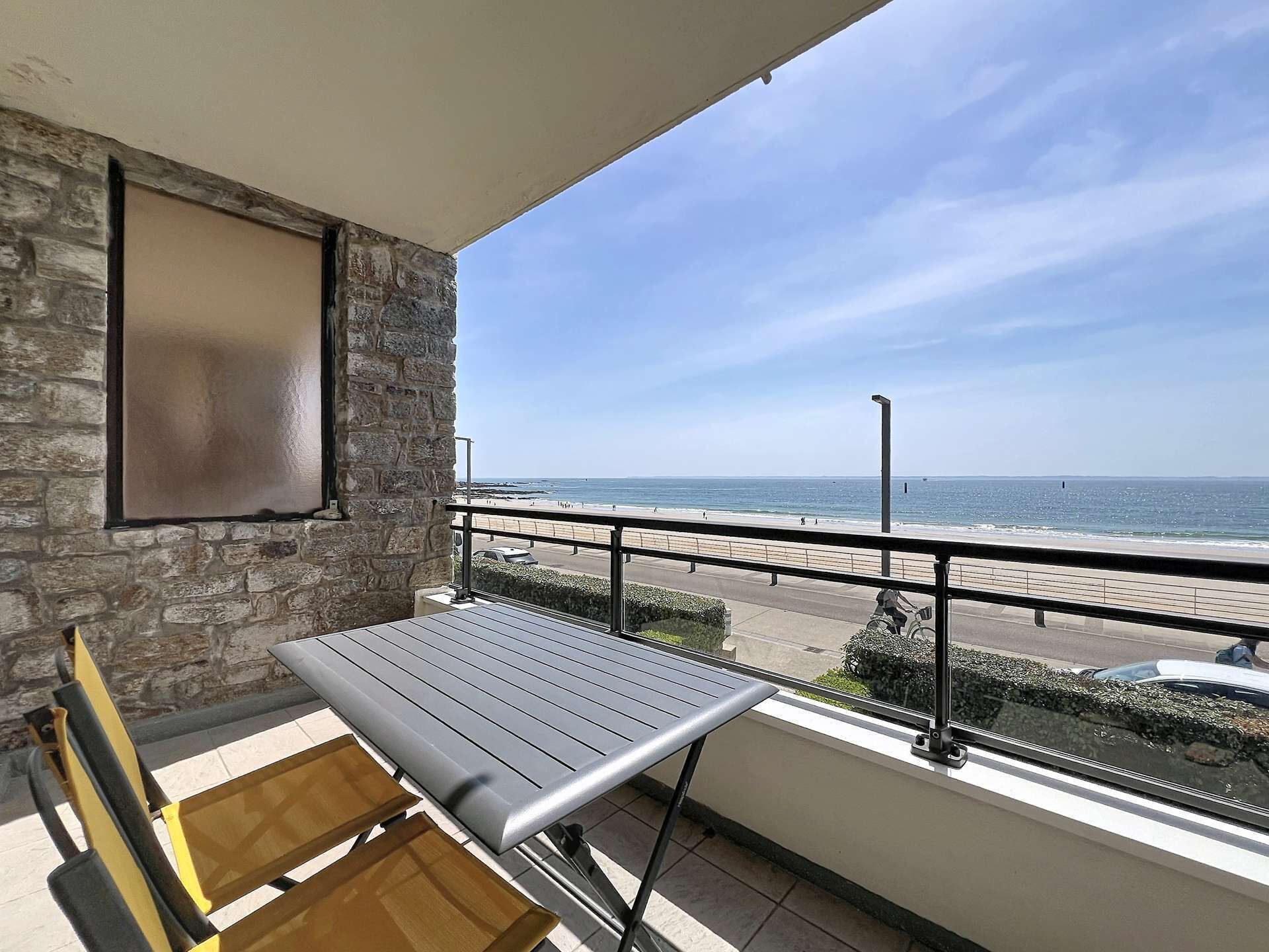 A balcony with a table and chairs, a stone wall, and a view of the beach and ocean.