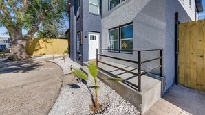 Une maison moderne à deux étages, avec une façade en briques grises, une clôture en bois et un petit palmier dans le jardin.