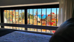 Ein Bett mit floralem Überwurf, ein Fenster mit Gitterstäben und Blick auf Gebäude und das Meer.