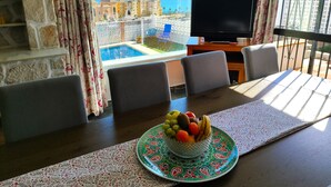 Ein Essbereich mit einem für zwei Personen gedeckten Tisch, einer Obstschale auf dem Tisch und Blick auf einen Pool sowie Gebäude im Freien.