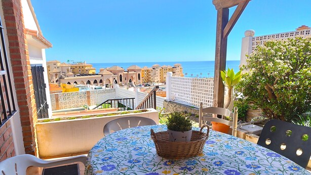 Ein Balkon mit Tisch für zwei Personen, einem Korbwicker-Korb mit einer Pflanze und Blick auf das Meer sowie Gebäude im Hintergrund.