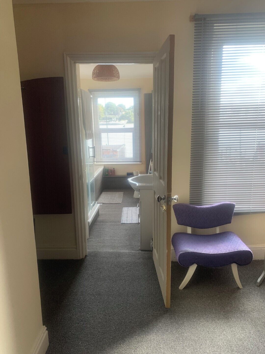 A narrow hallway with a door leading to a bathroom, a purple chair, and a window with blinds.