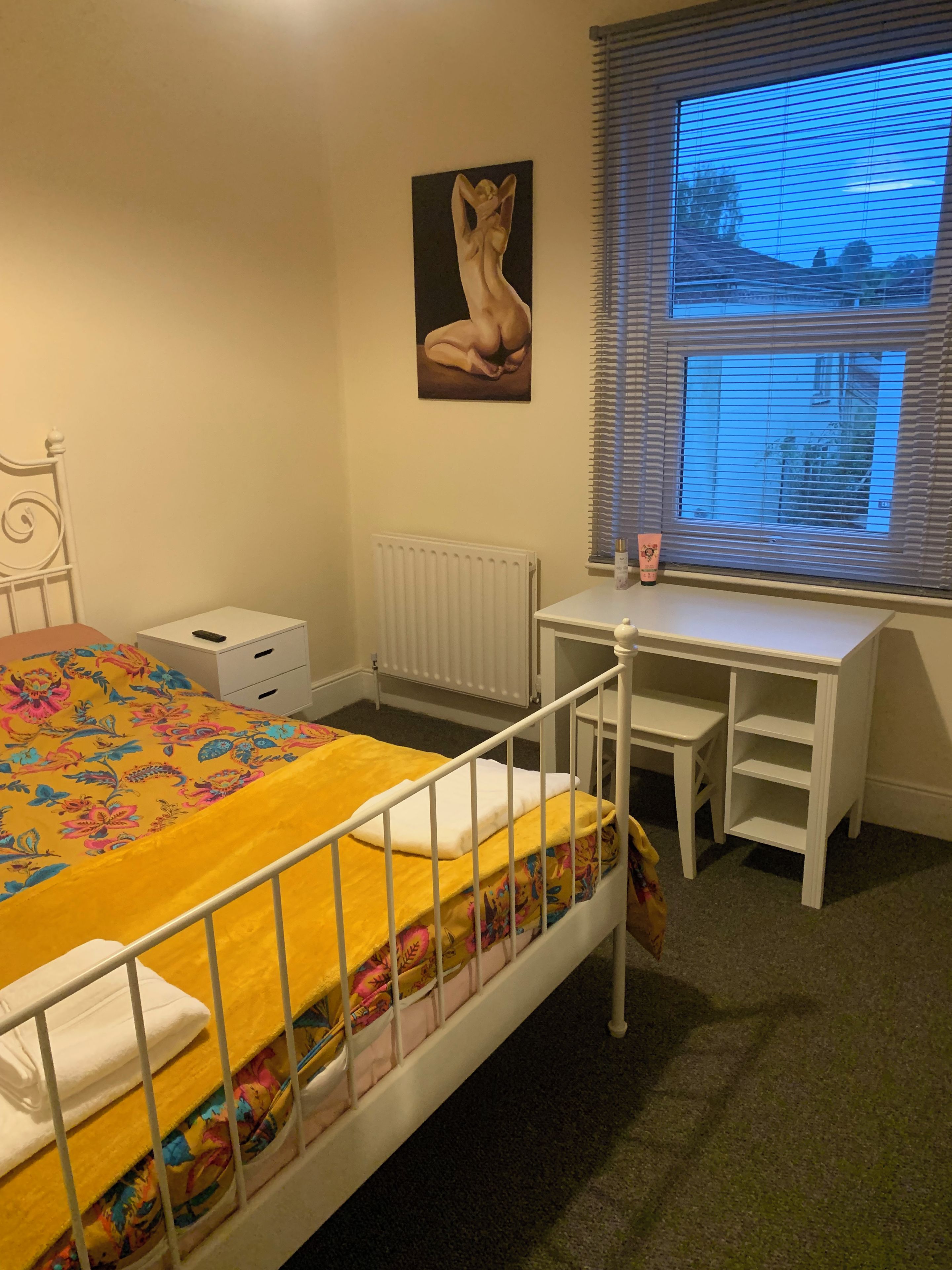 A bedroom with a bed, a desk, a radiator, and a window with blinds.