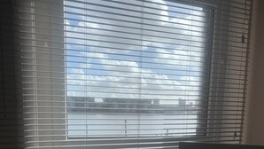 A room with a large window with horizontal blinds, a view of a body of water and buildings, and a partial view of a person in plaid pajama pants. - Perfect 2-bedroom house, Albert cottage with WiFi in charming Southampton (Southampton)