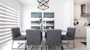 A modern dining area with a glass table and grey upholstered chairs, a geometric pendant light, and two abstract paintings on the wall. - Modern Executive Home-Sleeps12-KingBeds-FullHouse (Edmonton)
