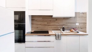 Uma cozinha moderna com armários brancos, forno embutido, micro-ondas, painel de madeira atrás do fogão, pia e bancada com uma garrafa e uvas.