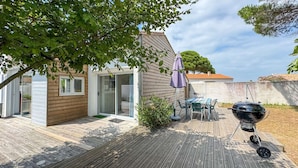 A wooden deck with a barbecue grill, a table with chairs, and a covered patio area. - Pretty single-storey villa with garden (LA COUARDE SUR MER)