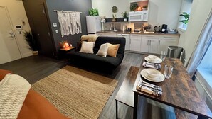 A modern living room with a black sofa, a wooden dining table set with plates and glasses, a kitchen area with white cabinets and a microwave, and a grey wall with a macrame decoration. - Boho Haven with Free Parking, Wifi and King Bed, Walk to Clubs, U of A and Food (Tucson)