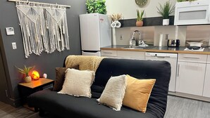 A modern living room with a black sofa, a macrame wall hanging, a kitchen area with white cabinets, and a microwave. - Boho Haven with Free Parking, Wifi and King Bed, Walk to Clubs, U of A and Food (Tucson)