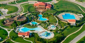 Vista aérea de un complejo turístico con múltiples piscinas, un edificio grande de techo rojo y jardines bien cuidados.